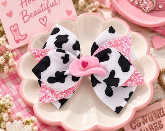 May include: A hair bow featuring a black and white cow print pattern layered with pink glitter fabric. The center of the bow is adorned with a small pink cowboy hat charm featuring a tiny red heart. The bow is displayed on a scalloped white plate.