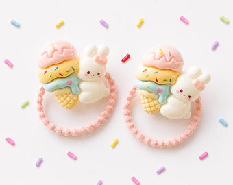 May include: Two hair ties featuring a three-scoop ice cream cone in pink, yellow, and blue, paired with a small white rabbit figure wearing a pink bow. The charms are attached to light pink elastic bands, surrounded by scattered colorful sprinkles.