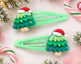 May include: Two light green metal hair clips, each featuring a three-dimensional Christmas tree ornament. The trees are green with small colorful dots, topped with a red and white Santa hat, and have a tan trunk at the base.