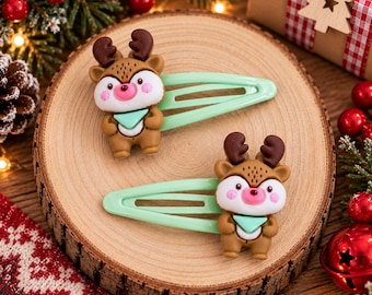 May include: Two hair clips featuring brown reindeer figures with dark brown antlers, pink cheeks, and light green neckerchiefs. Each reindeer is attached to a light mint green metal snap hair clip, showcasing a smooth finish and detailed facial features.