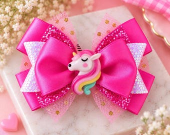 May include: A multi-layered hair bow featuring bright pink satin ribbon, glittery pink fabric, and sheer pink tulle with gold polka dots. A central unicorn charm with a pink, yellow, and teal mane is attached to the center of the bow.