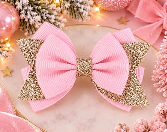 May include: A hair bow constructed from light pink ribbed fabric and gold glitter fabric. The bow features a central gold glitter knot, with layered pink loops and gold glitter pointed side accents, creating a multi-textured appearance.
