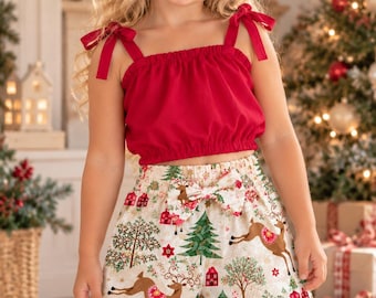 May include: A two-piece outfit consisting of a solid red sleeveless crop top with tie-shoulder straps and a pair of off-white shorts featuring a festive print of reindeer, evergreen trees, and small red houses with a decorative bow at the waist.