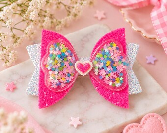 May include: A hair bow featuring hot pink textured fabric wings with clear heart-shaped pockets filled with multicolored star-shaped sequins. The bow includes a silver glitter base layer and a small pink heart charm with white dots at the center.