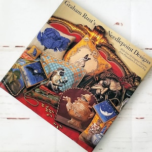 May include: A book titled "Graham Rust's Needlepoint Designs" with over 20 original patterns, from pincushion to seashell rug. The cover features a colorful illustration of needlepoint designs on a red velvet couch.