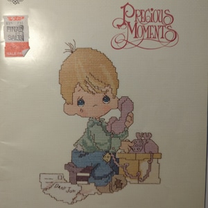 May include: A cross-stitch pattern featuring a Precious Moments figurine of a young boy on the phone. The boy is wearing a green shirt, blue pants, and a brown hat. He is sitting on a stool with a phone on a table beside him. The pattern is titled "Dear Jon" and is from the Precious Moments book PM-3.