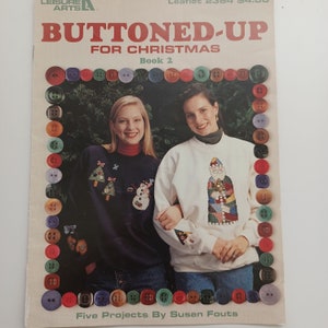 May include: A book cover with the title "Buttoned-Up for Christmas, Book 2" and a photo of two women wearing sweaters with Christmas-themed designs. The sweaters are decorated with buttons and the women are smiling. The book cover is surrounded by a border of colorful buttons.