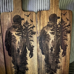 May include: Two rectangular wooden serving boards with handles, each featuring a dark laser-engraved illustration of a person wearing a cap and jacket, standing in a forest with pine trees, a deer, and birds flying in the sky.
