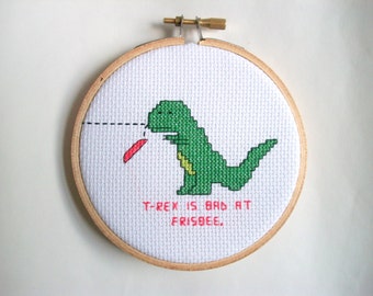 T-Rex is bad at flying discs, completed cross stitch with T-Rex being hit on the nose, in 4 in. natural wood embroidery hoop