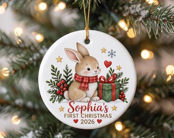 May include: A round white ceramic ornament featuring a tan bunny wearing a red plaid scarf next to a green gift box. The design includes red berries, green foliage, gold stars, and a snowflake. Text reads: Sophia's FIRST CHRISTMAS 2026.