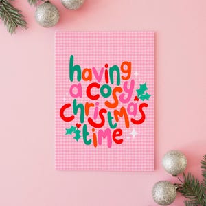 May include: A rectangular greeting card with a pink and white gingham pattern background. The text "having a cosy christmas time" is printed in a multi-colored, rounded font in shades of green, pink, red, and orange, accented by holly leaves.
