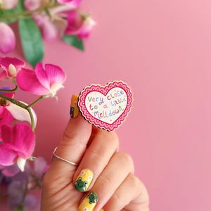 Pode incluir: Um broche de madeira em forma de cora&ccedil;&atilde;o com uma borda rosa ondulada e o texto "Very close to a little meltdown" em letras coloridas. O broche &eacute; segurado contra um fundo rosa, com flores rosas ao fundo.