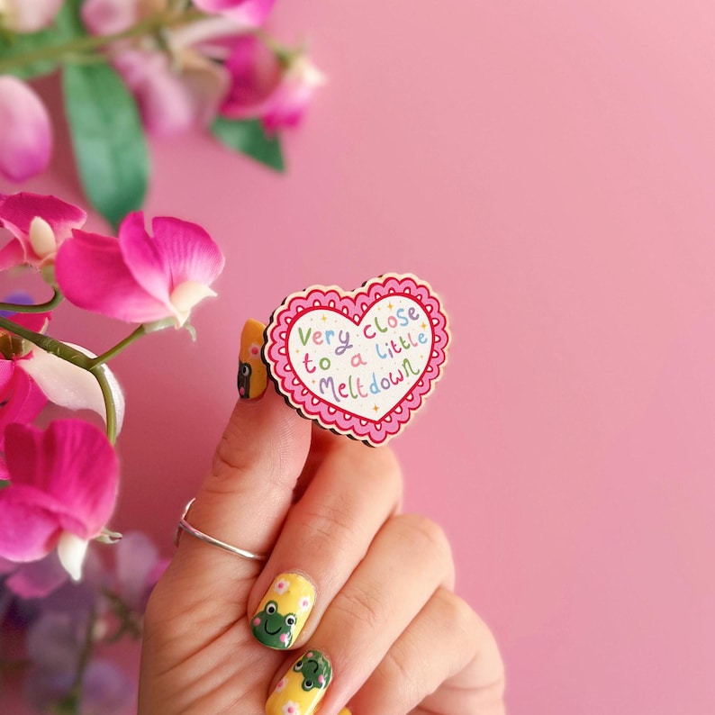 Pode incluir: Um broche de madeira em forma de cora&ccedil;&atilde;o com uma borda rosa ondulada e o texto "Very close to a little meltdown" em letras coloridas. O broche &eacute; segurado contra um fundo rosa, com flores rosas ao fundo.