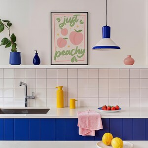 K&ouml;nnte beinhalten: Ein gerahmter Kunstdruck mit dem Schriftzug 'just peachy' in gr&uuml;ner Schreibschrift, illustriert mit rosa Pfirsichen und kleinen gelben Sternen auf hellrosa Hintergrund, in einer K&uuml;che mit blauen und gelben Akzenten.