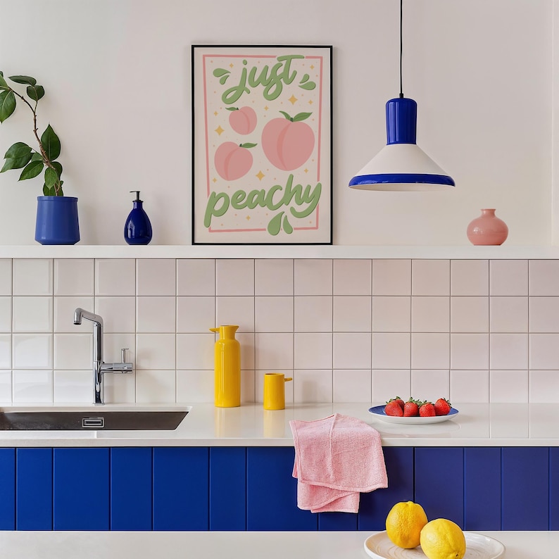 K&ouml;nnte beinhalten: Ein gerahmter Kunstdruck mit dem Schriftzug 'just peachy' in gr&uuml;ner Schreibschrift, illustriert mit rosa Pfirsichen und kleinen gelben Sternen auf hellrosa Hintergrund, in einer K&uuml;che mit blauen und gelben Akzenten.