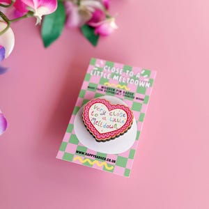 Pode incluir: Um distintivo de madeira em forma de cora&ccedil;&atilde;o com a frase "Very close to a little Meltdown". O pin &eacute; rosa, branco e castanho, e est&aacute; sobre um fundo xadrez verde e rosa. O site www.happybadger.co.uk &eacute; vis&iacute;vel.