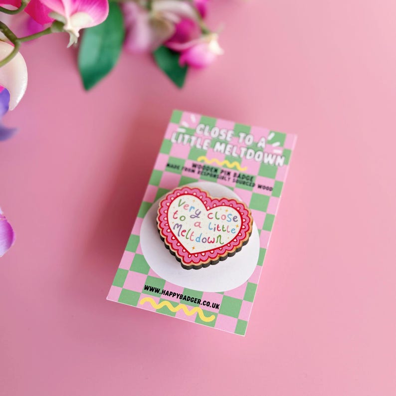 Pode incluir: Um distintivo de madeira em forma de cora&ccedil;&atilde;o com a frase "Very close to a little Meltdown". O pin &eacute; rosa, branco e castanho, e est&aacute; sobre um fundo xadrez verde e rosa. O site www.happybadger.co.uk &eacute; vis&iacute;vel.
