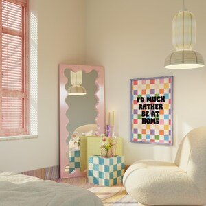 Puede incluir: Un dormitorio rosa con un sill&oacute;n blanco y esponjoso, un p&oacute;ster a cuadros de colores con el texto "I'd much rather be at home", un espejo ondulado rosa y una mesita auxiliar a cuadros con un jarr&oacute;n de flores.