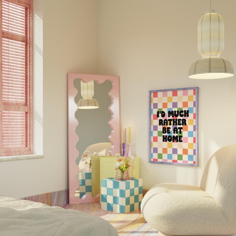 Puede incluir: Un dormitorio rosa con un sill&oacute;n blanco y esponjoso, un p&oacute;ster a cuadros de colores con el texto "I'd much rather be at home", un espejo ondulado rosa y una mesita auxiliar a cuadros con un jarr&oacute;n de flores.