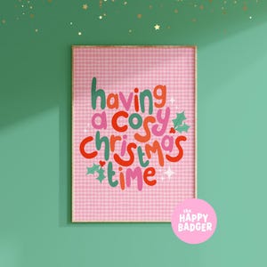May include: A rectangular wall art print featuring a pink and white gingham background with the text having a cosy christmas time in colorful bubble letters. The design includes small green holly leaves and white star accents throughout the print.