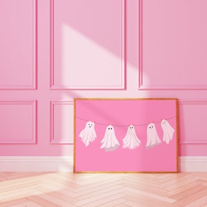 Pode incluir: Uma impress&atilde;o retangular emoldurada com fundo rosa, apresentando cinco ilustra&ccedil;&otilde;es de fantasmas brancos pendurados num fio preto com pequenos clipes laranja. Cada fantasma tem um padr&atilde;o distinto, incluindo bolinhas e detalhes em renda.