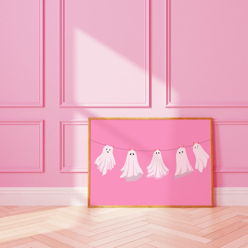 Pode incluir: Uma impress&atilde;o retangular emoldurada com fundo rosa, apresentando cinco ilustra&ccedil;&otilde;es de fantasmas brancos pendurados num fio preto com pequenos clipes laranja. Cada fantasma tem um padr&atilde;o distinto, incluindo bolinhas e detalhes em renda.