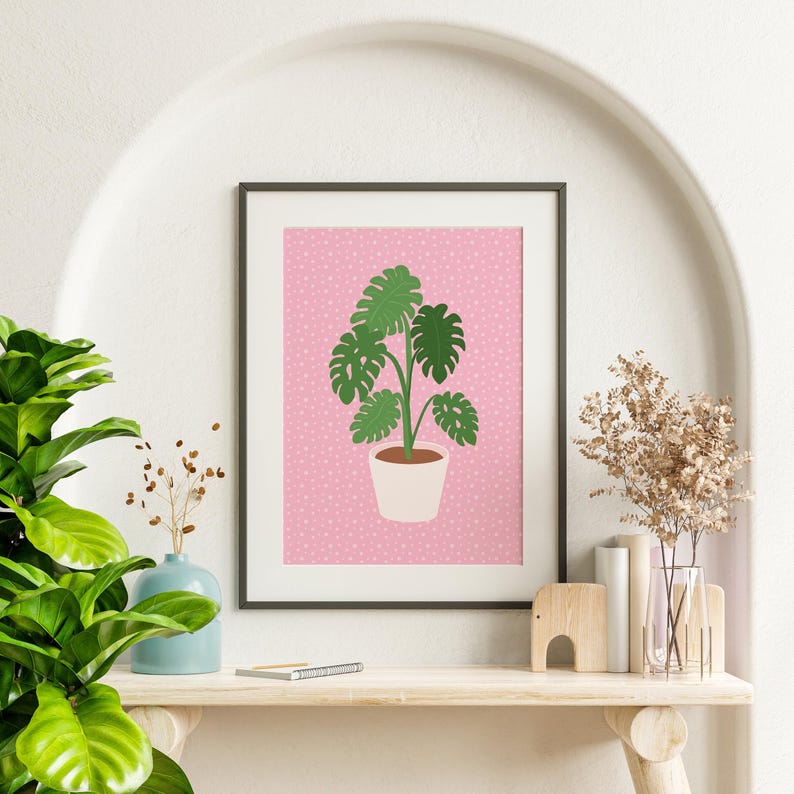 May include: A framed print of a green monstera plant in a white pot on a pink background with white polka dots.