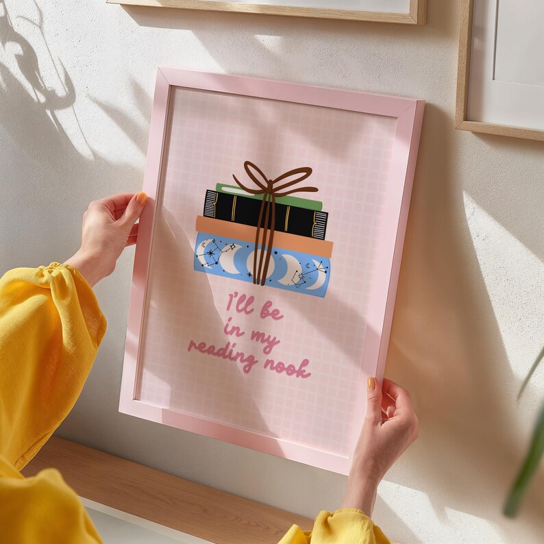 May include: A pink framed print with a stack of books tied with a brown ribbon. The books are decorated with moons and stars. The text on the print reads "I'll be in my reading nook."