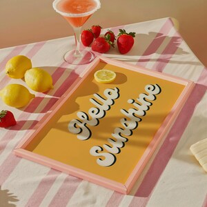 May include: A pink framed tray with a yellow background and the text "Hello Sunshine" in white. The tray is on a pink and white striped surface with lemons and strawberries.