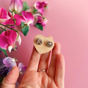 Pode incluir: Dois bot&otilde;es de lapela prateados em um cart&atilde;o em forma de cora&ccedil;&atilde;o, marrom claro. O cart&atilde;o &eacute; segurado em uma m&atilde;o, com um fundo rosa e flores rosa e roxas no canto superior esquerdo. Os pinos t&ecirc;m um acabamento met&aacute;lico brilhante.