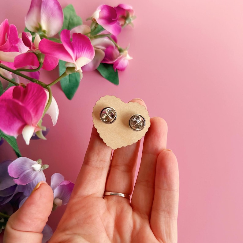 Pode incluir: Dois bot&otilde;es de lapela prateados em um cart&atilde;o em forma de cora&ccedil;&atilde;o, marrom claro. O cart&atilde;o &eacute; segurado em uma m&atilde;o, com um fundo rosa e flores rosa e roxas no canto superior esquerdo. Os pinos t&ecirc;m um acabamento met&aacute;lico brilhante.
