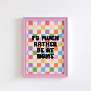 Puede incluir: Una impresi&oacute;n enmarcada en rosa con un fondo a cuadros de colores y el texto "I'd much rather be at home" en negro.