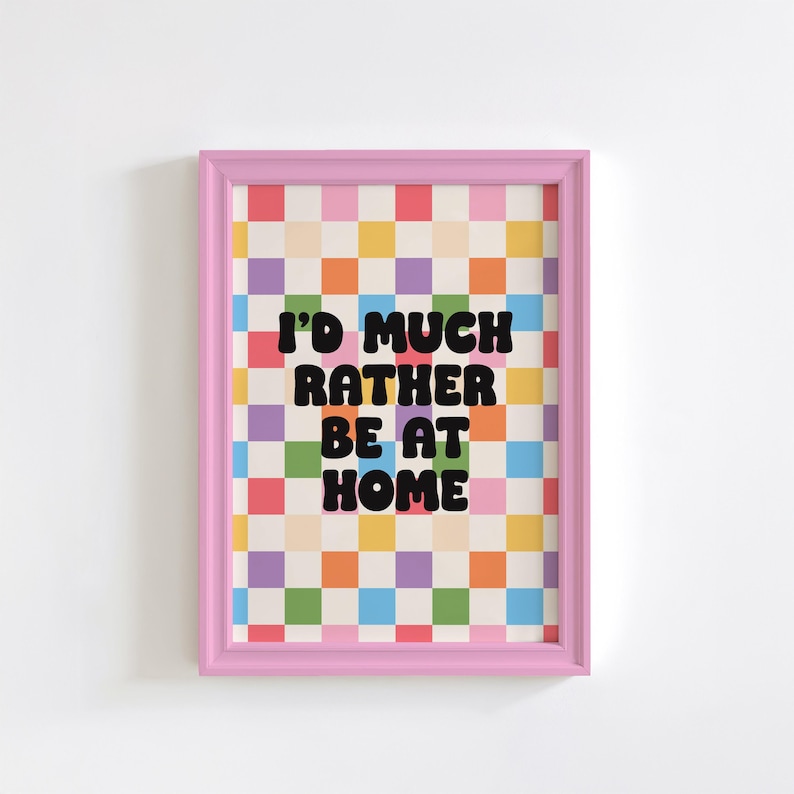 Puede incluir: Una impresi&oacute;n enmarcada en rosa con un fondo a cuadros de colores y el texto "I'd much rather be at home" en negro.