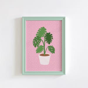 May include: A framed print of a green monstera plant in a white pot on a pink background with white polka dots.