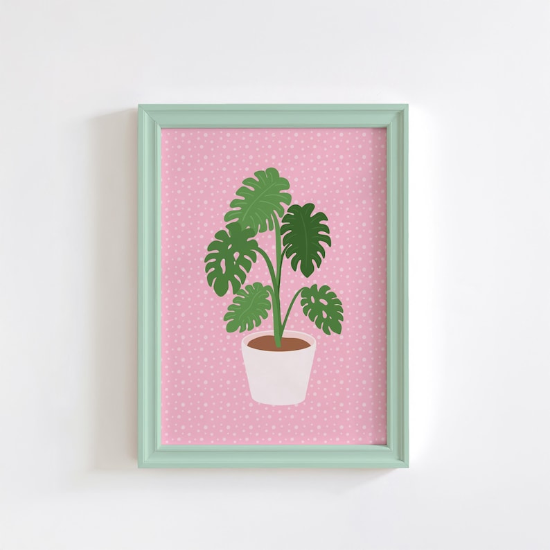 May include: A framed print of a green monstera plant in a white pot on a pink background with white polka dots.