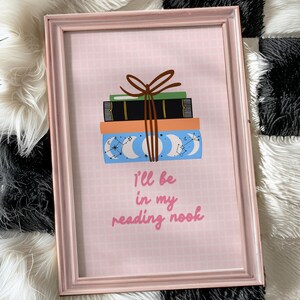 May include: A pink framed print with a stack of books tied with a brown ribbon. The books have a blue and white celestial pattern. The text on the print reads "I'll be in my reading nook."