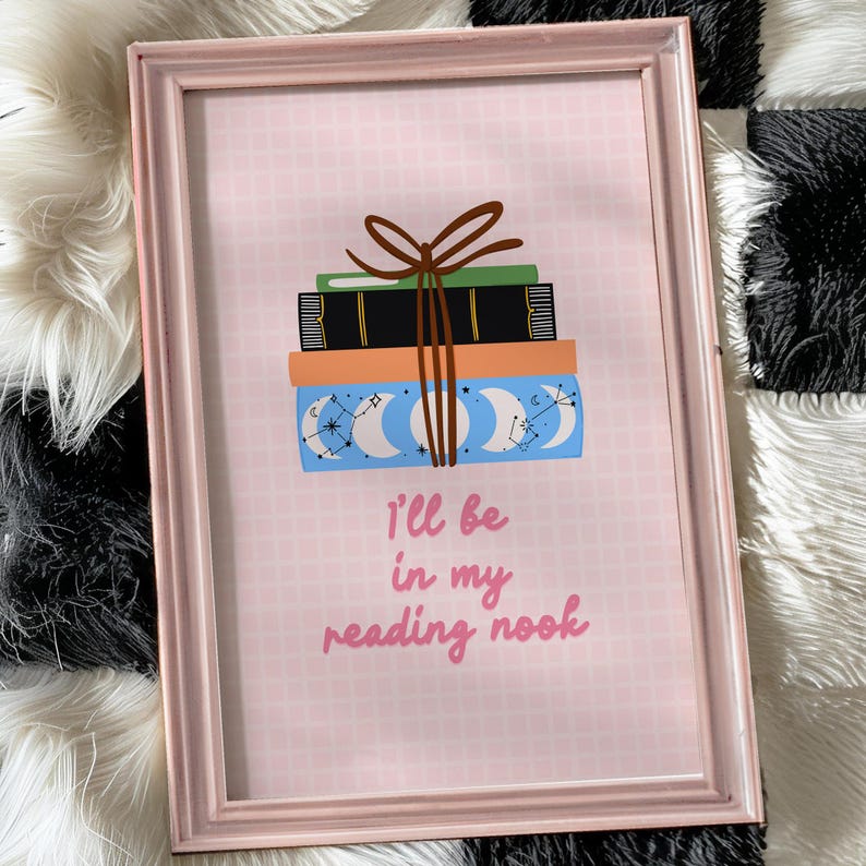 May include: A pink framed print with a stack of books tied with a brown ribbon. The books have a blue and white celestial pattern. The text on the print reads "I'll be in my reading nook."