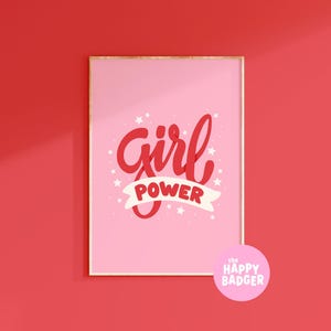 Può includere: Una stampa da parete rettangolare incorniciata con sfondo rosa chiaro, il testo rosso "Girl" in corsivo e il testo rosso "POWER" in un carattere sans-serif in grassetto su un nastro bianco, circondato da piccole stelle bianche.