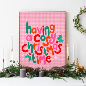 May include: A rectangular wall art print in a gold frame featuring a pink gingham background with the text "having a cosy christmas time" in colorful, rounded, bubbly lettering, accented by small green holly leaves and white star shapes.