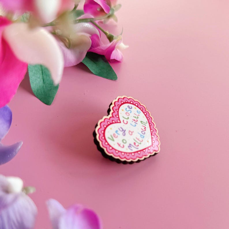 Pode incluir: Um alfinete em forma de cora&ccedil;&atilde;o com uma borda ondulada e a frase "Very close to a little meltdown" em letras coloridas. O alfinete &eacute; rosa e branco com uma borda preta, sobre um fundo rosa com flores.