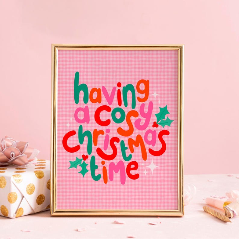 May include: A framed wall art print featuring a pink gingham background with the text "having a cosy christmas time" in colorful, rounded lettering. The design includes green holly leaves with red berries and small white star accents.