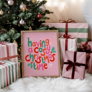 May include: A rectangular framed print with a light pink gingham background featuring the text having a cosy christmas time in red, green, and pink lettering, accented with small green holly leaves and white star shapes on the print surface.
