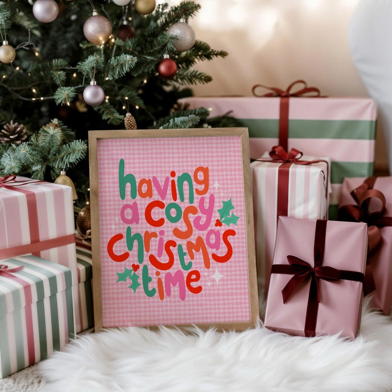 May include: A rectangular framed print with a light pink gingham background featuring the text having a cosy christmas time in red, green, and pink lettering, accented with small green holly leaves and white star shapes on the print surface.
