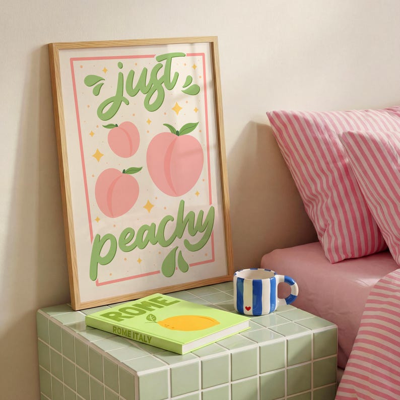 K&ouml;nnte beinhalten: Ein gerahmter Kunstdruck mit dem Schriftzug "just peachy" in gr&uuml;ner Schreibschrift, erg&auml;nzt durch drei rosa Pfirsichillustrationen und kleine gelbe Sterne. Der Druck befindet sich in einem hellen Holzrahmen auf einer gefliesten Fl&auml;che.