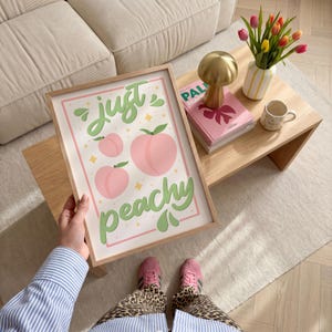 K&ouml;nnte beinhalten: Ein rechteckiger gerahmter Kunstdruck mit wei&szlig;em Hintergrund, drei hellrosa Pfirsich-Illustrationen und dem gr&uuml;nen Schriftzug "just peachy" in Schreibschrift. Das Design enth&auml;lt kleine gelbe Sternakzente und einen d&uuml;nnen rosa Rand.