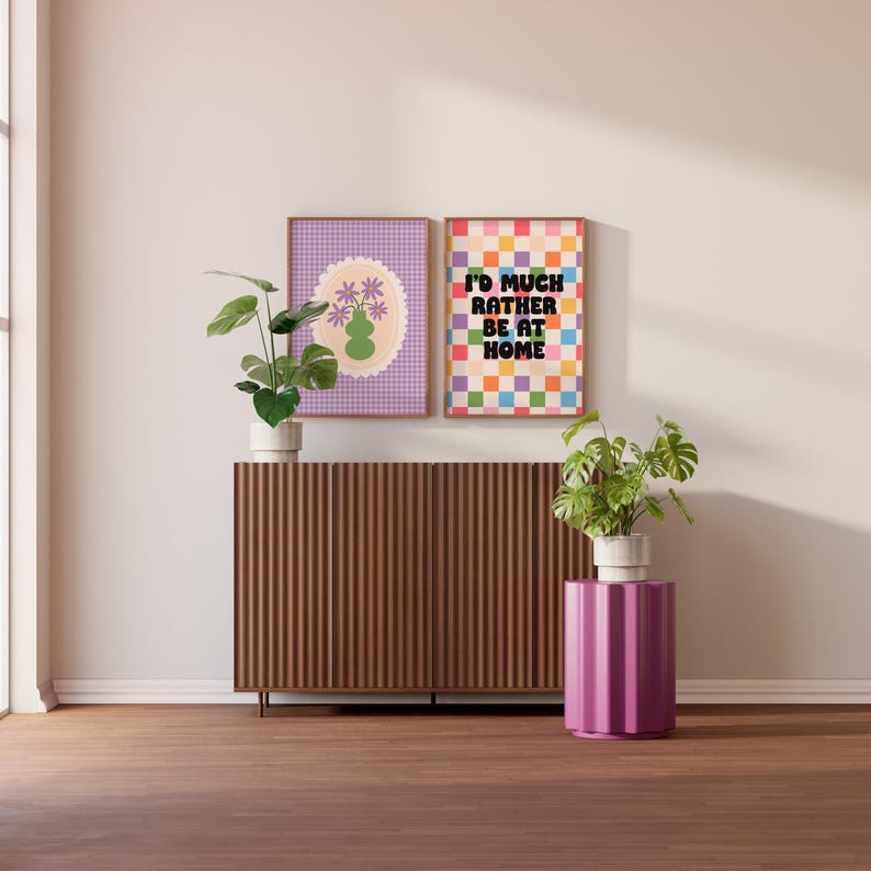 Puede incluir: Dos impresiones enmarcadas en un gabinete de madera con una mesa auxiliar morada. La impresi&oacute;n de la izquierda tiene un fondo de cuadros morados y blancos con un jarr&oacute;n verde y flores moradas. La impresi&oacute;n de la derecha tiene un fondo a cuadros de colores con el texto "I'd much rather be at home".