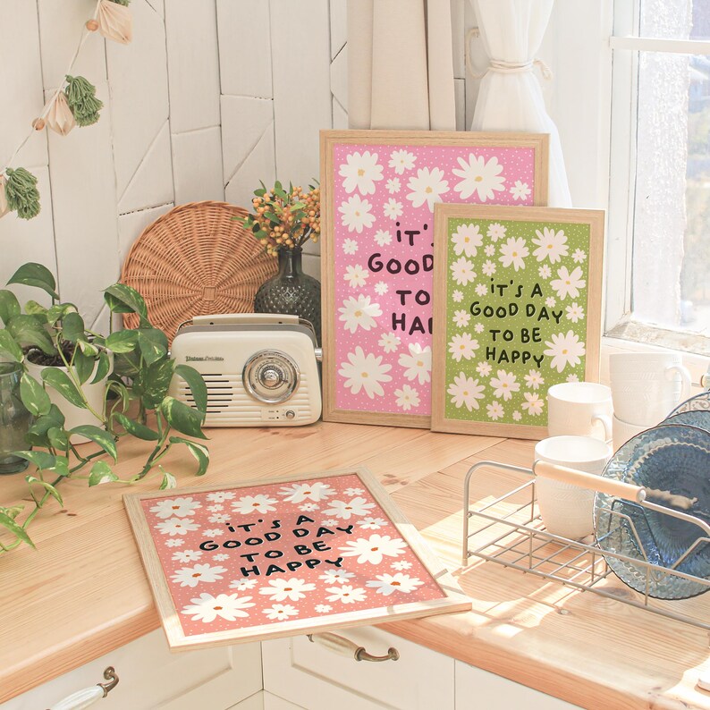 Op de afbeelding: Drie ingelijste prints met een madeliefjespatroon en de tekst "It's a good day to be happy" in zwarte letters. De prints staan op een houten oppervlak met een radio, een plant en een afdruiprek.