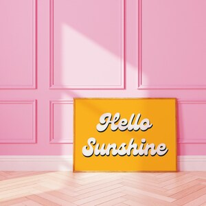 May include: A yellow framed poster with the text "Hello Sunshine" in white cursive lettering against a yellow background. The poster is leaning against a pink wall with white trim.