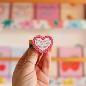 Pode incluir: Um anel em forma de cora&ccedil;&atilde;o com uma borda ondulada e o texto "Very close to a little Meltdown" em letras coloridas. O anel &eacute; segurado por uma m&atilde;o com um anel de prata e uma unha amarela com um desenho de desenho animado.
