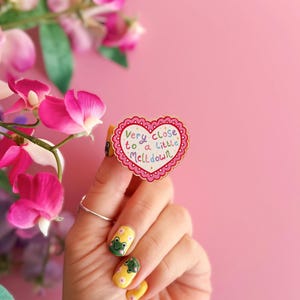 Pode incluir: Um alfinete em forma de cora&ccedil;&atilde;o com uma borda ondulada e o texto "Very close to a little meltdown" em letras coloridas. O alfinete &eacute; rosa e branco, e &eacute; segurado por uma m&atilde;o com esmalte de unha amarelo e desenhos de sapos. Flores cor-de-rosa no fundo.